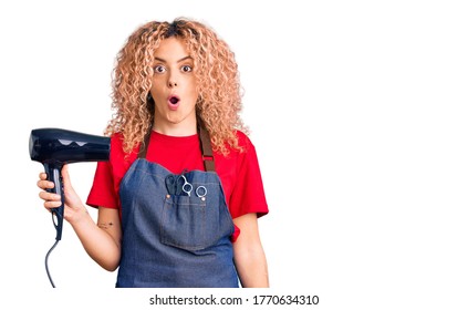 Young Blonde Woman With Curly Hair Wearing Hairdresser Apron And Holding Dryer Blow Scared And Amazed With Open Mouth For Surprise, Disbelief Face 