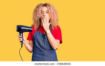 Young Blonde Woman With Curly Hair Wearing Hairdresser Apron And Holding Dryer Blow Covering Mouth With Hand, Shocked And Afraid For Mistake. Surprised Expression 