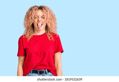 Young Blonde Woman With Curly Hair Wearing Casual Red Tshirt Sticking Tongue Out Happy With Funny Expression. Emotion Concept. 