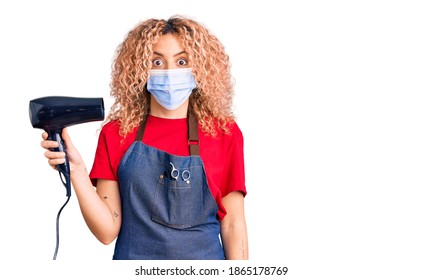 Young Blonde Woman With Curly Hair Holding Dryer Blow Wearing Safety Mask For Coranvirus Scared And Amazed With Open Mouth For Surprise, Disbelief Face 