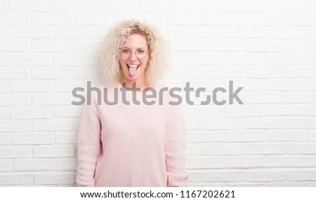 Young blonde woman with curly hair over white brick wall sticking tongue out happy with funny expression. Emotion concept.