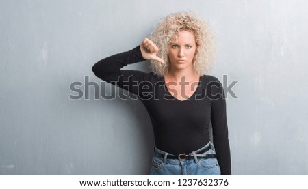 Young blonde woman with curly hair over grunge grey background with angry face, negative sign showing dislike with thumbs down, rejection concept