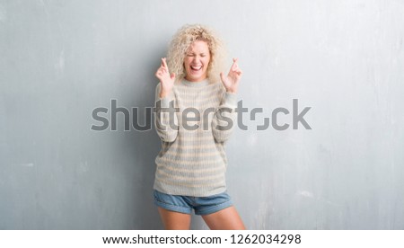 Young blonde woman with curly hair over grunge grey background smiling crossing fingers with hope and eyes closed. Luck and superstitious concept.