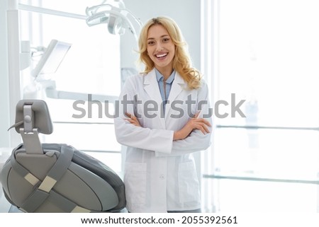 Young blonde woman dentist posing at modern dental cabinet, standing next to dental chair with arms crossed on chest and cheerfully smiling at camera, copy space. Pretty stomatologist at dental clinic