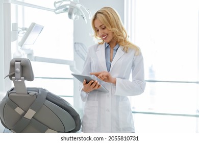 Young blonde woman dentist standing by dental chair, using digital tablet at newest modern dental clinic, chatting with client or checking her schedule online, copy space. Medical appointment concept - Powered by Shutterstock