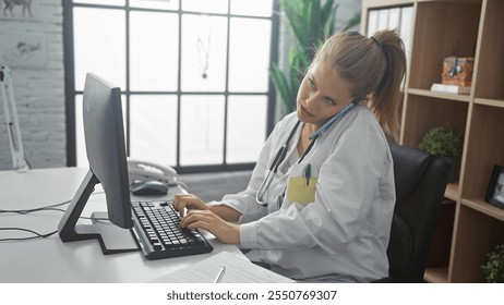 Young blonde woman, a doctor, multitasks on a computer while making a phone call in a bright hospital office - Powered by Shutterstock