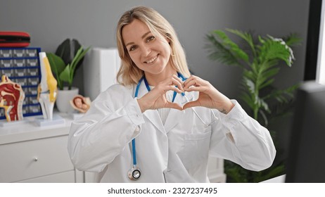 Young blonde woman doctor smiling doing heart gesture with hands at clinic