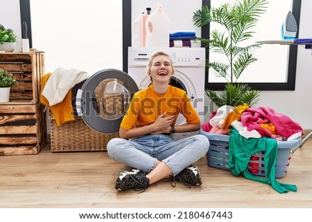 Young blonde woman doing laundry sitting by washing machine smiling and laughing hard out loud because funny crazy joke with hands on body. 