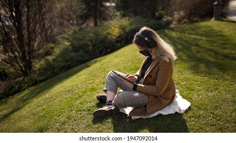 young blonde woman dressed in a beige coat in a black mask sitting on the grass with handsfree or wireless headphones on searching something in the tablet