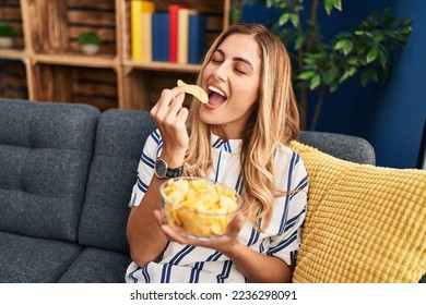 Young blonde woman eating chips potatoes sitting on sofa at home