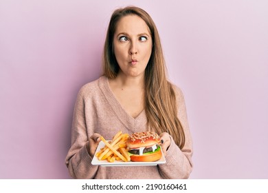 Young blonde woman eating a tasty classic burger with fries making fish face with mouth and squinting eyes, crazy and comical.  - Shutterstock ID 2190160461