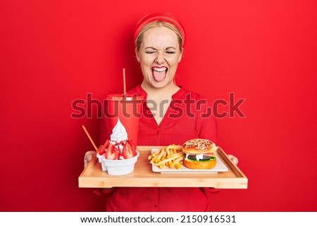 Young blonde woman eating a tasty classic burger with fries and soda sticking tongue out happy with funny expression. 