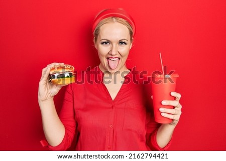 Young blonde woman eating a tasty classic burger with fries and soda sticking tongue out happy with funny expression. 