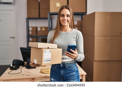 Young blonde woman ecommerce business worker using smartphone holding packages at office