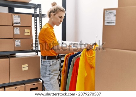 Young blonde woman ecommerce business worker organizing clothes on coat rack at office
