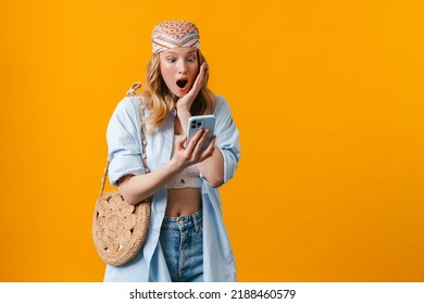 Young blonde woman expressing surprise and using cellphone isolated over yellow background