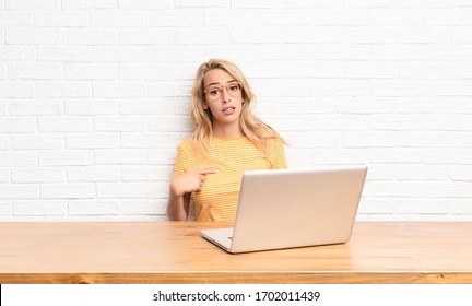 young blonde woman feeling confused, puzzled and insecure, pointing to self wondering and asking who, me? using a laptop