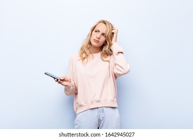 Young Blonde Woman Feeling Puzzled And Confused, Scratching Head And Looking To The Side Using A Smartphone