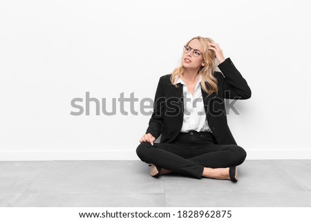 young blonde woman feeling puzzled and confused, scratching head and looking to the side sitting on the floor
