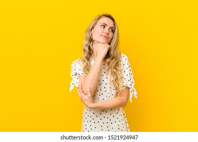 young blonde woman feeling thoughtful, wondering or imagining ideas, daydreaming and looking up to copy space against yellow wall - Shutterstock ID 1521924947