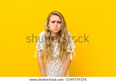 young blonde woman with a goofy, crazy, surprised expression, puffing cheeks, feeling stuffed, fat and full of food against yellow wall