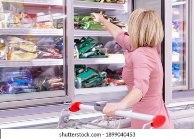 A Young Blonde Woman In A Grocery Store In The Frozen Food Department Selects A Product. Back View.