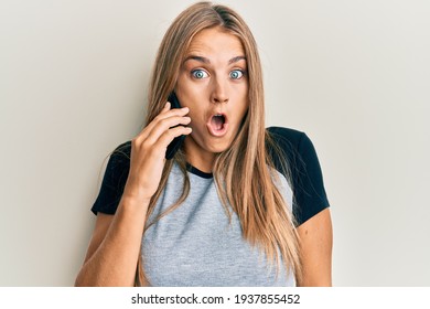Young blonde woman having conversation talking on the smartphone scared and amazed with open mouth for surprise, disbelief face 