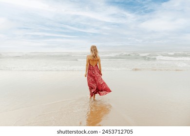 Young blonde woman with her back turned dressed in summer dress walking alone along the shore of the beach towards the sea. - Powered by Shutterstock