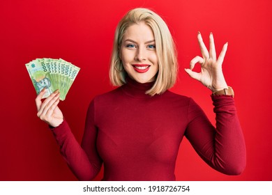 Young blonde woman holding 1000 chilean pesos doing ok sign with fingers, smiling friendly gesturing excellent symbol 