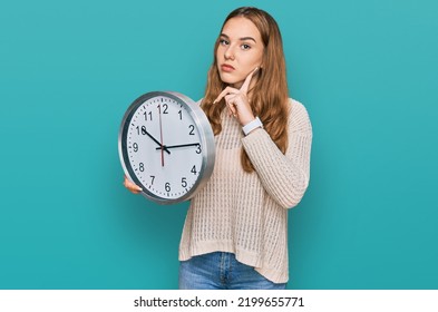 Young Blonde Woman Holding Big Clock Serious Face Thinking About Question With Hand On Chin, Thoughtful About Confusing Idea 