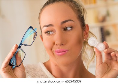 Young blonde woman holding contact lens case on hand and holding in her other hand a blue glasses on blurred background., eyesight and eyecare concept