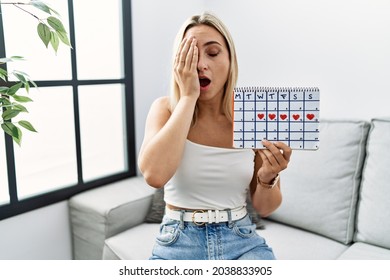 Young blonde woman holding heart calendar yawning tired covering half face, eye and mouth with hand. face hurts in pain. 