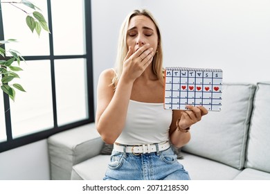 Young blonde woman holding heart calendar bored yawning tired covering mouth with hand. restless and sleepiness. 