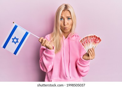 Young Blonde Woman Holding Israel Flag And Shekels Banknotes Depressed And Worry For Distress, Crying Angry And Afraid. Sad Expression. 