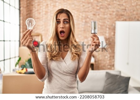 Young blonde woman holding led lightbulb and incandescent bulb in shock face, looking skeptical and sarcastic, surprised with open mouth 