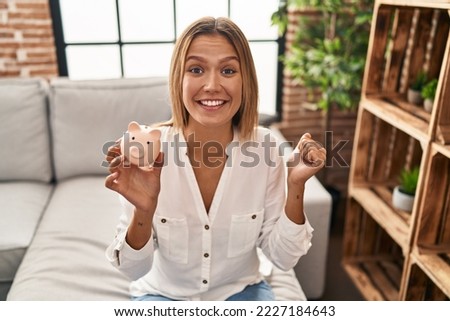 Young blonde woman holding piggy bank screaming proud, celebrating victory and success very excited with raised arm 
