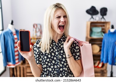 Young Blonde Woman Holding Shopping Bags Showing Smartphone Screen Angry And Mad Screaming Frustrated And Furious, Shouting With Anger. Rage And Aggressive Concept. 