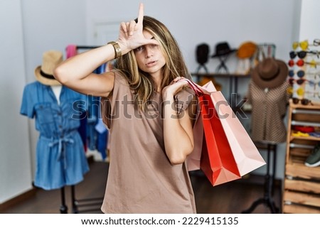 Young blonde woman holding shopping bags at retail shop making fun of people with fingers on forehead doing loser gesture mocking and insulting. 