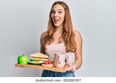 Young blonde woman holding tray with breakfast food celebrating crazy and amazed for success with open eyes screaming excited. 