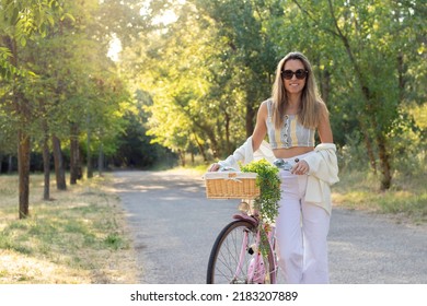 Young Blonde Woman Holding A Vintage Pink Bicycle With Wicker Basket And Plant Hanging In Nature Enviroment