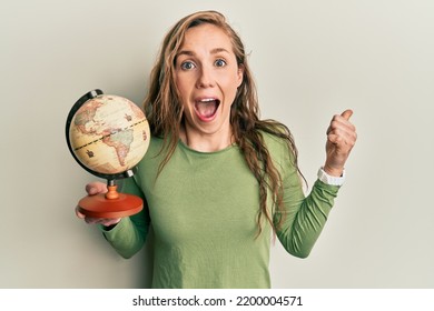 Young Blonde Woman Holding Vintage World Ball Pointing Thumb Up To The Side Smiling Happy With Open Mouth 