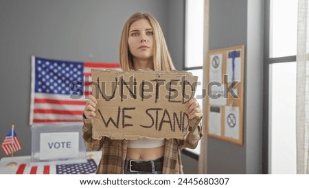 A young blonde woman holds a 'united we stand' sign indoors with the american flag and voting booth, symbolizing us electoral participation.
