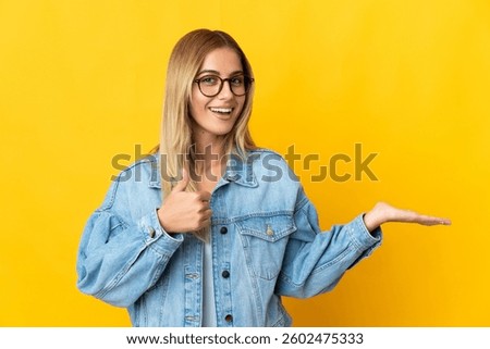 Young blonde woman isolated on yellow background holding copyspace imaginary on the palm to insert an ad and with thumbs up
