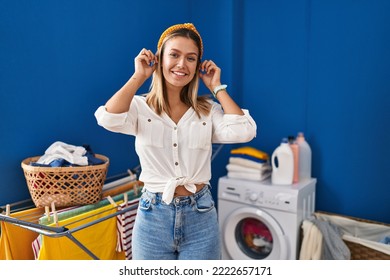 Young Blonde Woman At Laundry Room Smiling Pulling Ears With Fingers, Funny Gesture. Audition Problem 