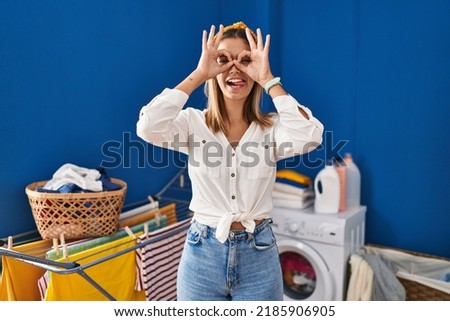 Young blonde woman at laundry room doing ok gesture like binoculars sticking tongue out, eyes looking through fingers. crazy expression. 