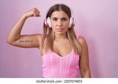 Young Blonde Woman Listening To Music Using Headphones Strong Person Showing Arm Muscle, Confident And Proud Of Power 