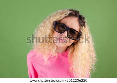 Young blonde woman with long curly hair in pink sweater on green background in 3d  cinema glasses serious focused look  to camera