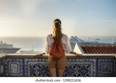 Young blonde woman looking at city view with sea from decorated balcony in azulejo tiles in Lisbon, Portugal
