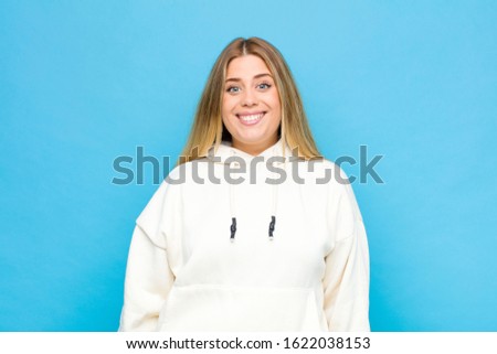 young blonde woman looking happy and goofy with a broad, fun, loony smile and eyes wide open against flat wall