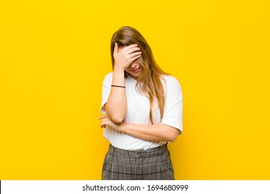 young blonde woman looking stressed, ashamed or upset, with a headache, covering face with hand against orange wall